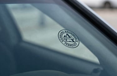A macro photograph focused on the official safety certification stamp on the corner of a high-quality auto glass pane. The glass reflects a clean, professional North American environment with cool steel blue and ice blue lighting.