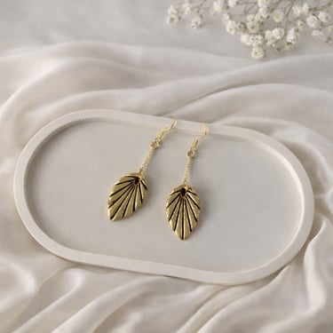 Elegant gold leaf dangle earrings resting on a white ceramic jewelry tray with silk fabric.