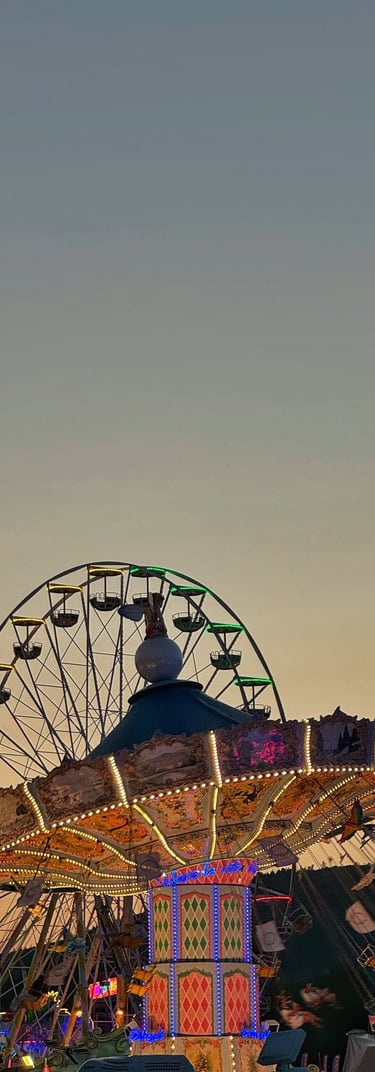 Beleuchtete Karussells, dahinter ein Riesenrad und ein Schaukelkarussell, in der Abenddämmerung