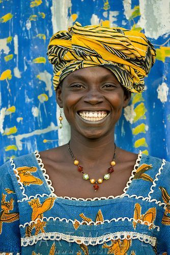 Smiling woman wearing a yellow African headwrap and patterned blue dress against a blue background.