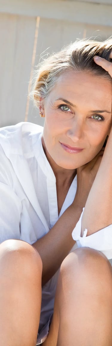 A blonde woman with green eyes wearing a white linen shirt posing on a sunny deck.