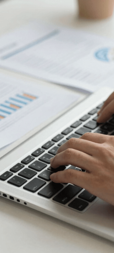 A close-up of a person typing on a laptop keyboard surrounded by financial audit reports in a Global / Corporate office setting. Well-lit, off-white desk.