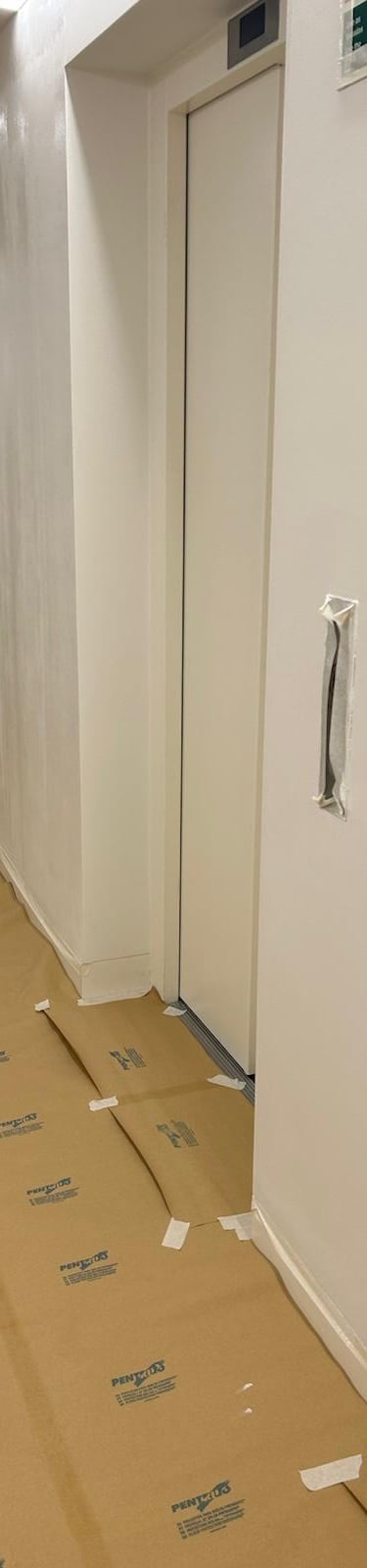 Professional painter applying white paint to a hallway wall with protected flooring.