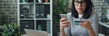 Eine Frau hat ihre Bankkarte in der Hand, sitzt vor einem Schreibtisch mit ihrem Laptop darauf. 