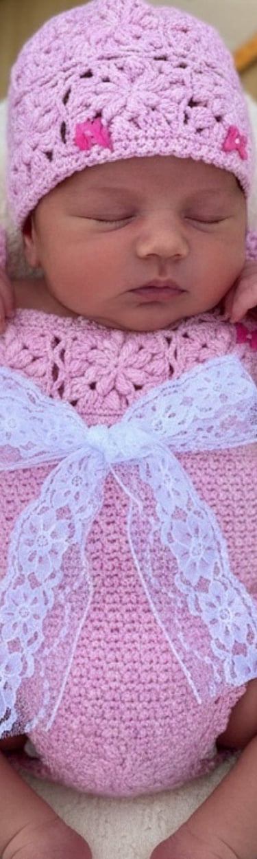 Sleeping newborn baby girl wearing a pink crochet romper and matching hat with white lace bow.