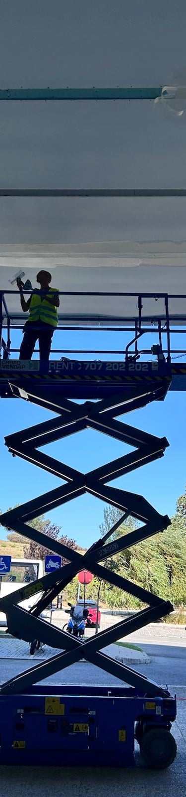 A construction worker on a blue scissor lift platform performing ceiling maintenance at a modern building entrance.