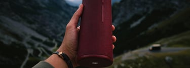 a person holding a red bluetooth speaker in their hand in the mountains