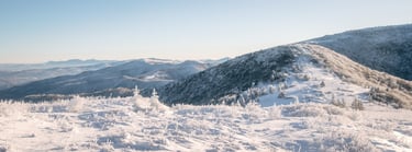 Panoramic winter landscape of snow-covered mountain peaks and frost-covered trees under a clear blue sky.