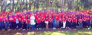 A large group of people wearing red LEGO t-shirts posing for a panoramic photo in a wooded park.