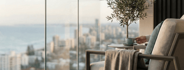 Luxury modern balcony with glass railings, a lounge chair, and a scenic coastal city view at sunset.