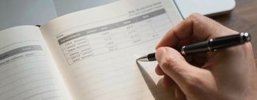 Close-up overhead view of an open notebook on a desk, a hand writing with a pen alongside a printed salary comparison sheet, natural window light casting soft shadows, a laptop keyboard visible at the top edge of the frame.
