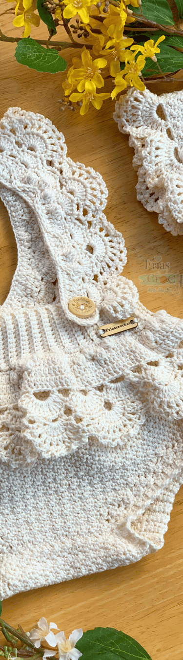 Handmade cream crochet baby romper with ruffled straps and matching bonnet on a wooden surface.