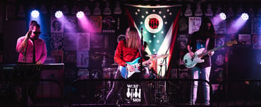 A live rock band performs on a stage with neon lighting and a poster-filled backdrop.