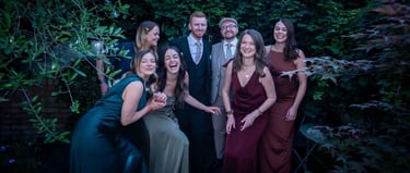 A group of friends in formal wedding attire laughing together in a lush green garden setting.