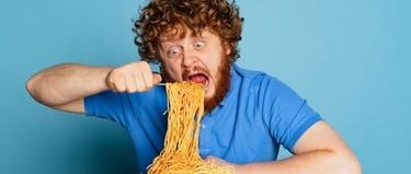 A hungry man with curly red hair eating a large plate of spaghetti noodles against a blue background.