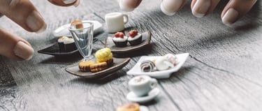 A person with manicured fingernails arranging realistic miniature food items and tiny dishes on a wooden surface.