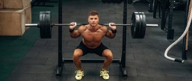 Muscular man performing a heavy barbell back squat in a gym setting for strength training.