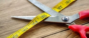 Red handled sewing scissors cutting a yellow measuring tape on a wooden table.