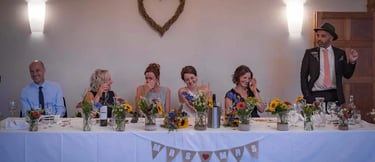 Wedding guests laugh during speeches at a rustic head table decorated with sunflowers and a wicker heart.