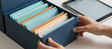 Close-up of hands organizing color-coded folders in a tidy North American / US home office. A tablet rests nearby showing a clear checklist. The color palette features dark slate blue folders and soft aqua tabs against an off-white desk, symbolizing clarity and organization.