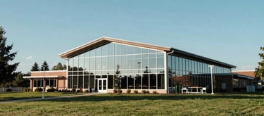 A wide-angle landscape photograph of a modern community center with glass walls reflecting a clear blue North American sky, surrounded by parkland, conveying quiet strength and stability.