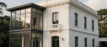 Exterior shot of a colonial-style building with modern glass expansions in a lush South American garden. The architecture is refined and elegant, surrounded by native flora under a soft blue sky. Palette: off-white and warm grey.