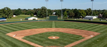 A wide-angle landscape shot of a beautifully maintained softball complex in Indiana, US, featuring multiple fields with green grass and clean white foul lines under a clear blue sky.