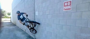 Rider performing a wall ride on a BMX bike against a concrete block wall next to a No Parking sign c