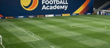 A wide shot of a state-of-the-art soccer facility in France with multiple green pitches, featuring a large banner with the Total Football Academy logo in yellow orange and white.