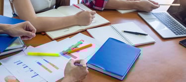 a group of people studying at a table with notebooks, pens, papers and a laptop
