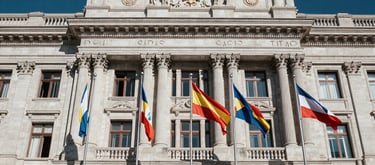 A long shot of an impressive stone institutional building with several international flags flying out front, majestic and authoritative style, global Spanish-speaking context, colors of dark blue and off-white.