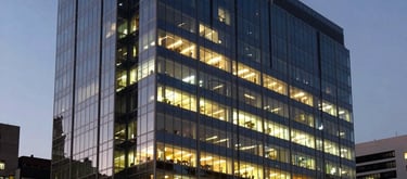 The sleek exterior of a modern glass office building in a prestigious South American district at dusk, glowing with warm internal light.