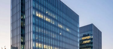 Wide-angle architectural photography of a modern, sleek biotechnology research building at sunset in a North American / International technology hub. The glass facade reflects a palette of deep blue and soft cyan, with warm interior laboratory lights visible.