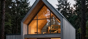 Exterior photography of a modern spiritual sanctuary in the North American Pacific Northwest. The building features light grey wood and glass, nestled among tall pines at dusk. Warm light glows from within.