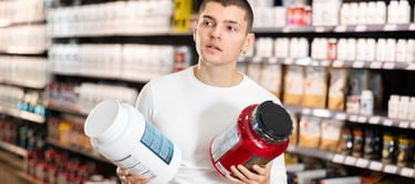 A man in a sports nutrition store comparing two large tubs of protein powder supplements.