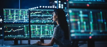 Female financial analyst monitoring stock market data and real-time trading charts on multiple computer monitors.