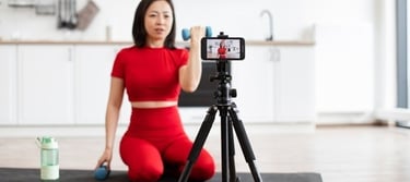 A fitness influencer filming a dumbbell arm workout at home using a smartphone on a tripod.