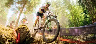 Professional mountain biker racing down a rocky forest trail during a cross-country MTB competition.