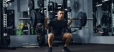 A fit man performing a heavy barbell back squat in a modern gym with weights and racks.