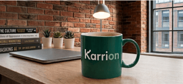 A close up of a Karrion Consulting coffee mug on a table with a laptop and books in the background.