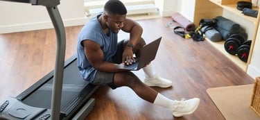 A man in gym clothes sits on a treadmill using a laptop for his home workout routine.