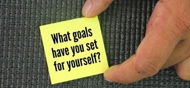 Hand holding a yellow sticky note with goal setting motivation text on a grey textured background.