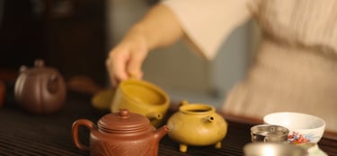 Person pouring water from a teapot with cups and a tea strainer on a wooden table