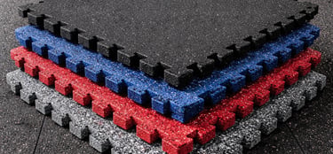 Stacked interlocking rubber gym floor tiles in black, blue, red, and grey inside a fitness center.