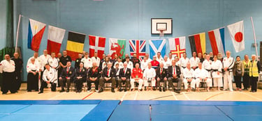 A group of martial arts students and instructors in white gi pose before international flags at a dojo.