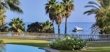 Luxury villa in Marbella overlooking a yacht on the blue ocean with palm trees.