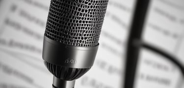 A person is speaking into a microphone, with a blurred background suggesting the setting is a recording or broadcast studio. The lighting is warm, creating a focused and professional atmosphere. A computer screen with editing software is partially visible in the background.