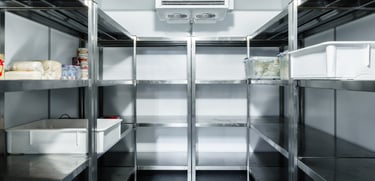 Stainless steel shelves line the walls of a commercial cold room, holding plastic containers, food items, and drinks. The roo