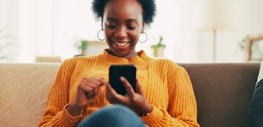 Smiling young Black woman in a yellow sweater using a smartphone while relaxing on a sofa.