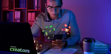 Homem em um escritório com luzes neon mexendo no notebook e celular. Há símbolos de likes e dinheiro